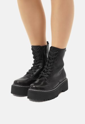 Botas de tobillo negras con cordones, acabado brillante, suela de plataforma gruesa y costuras en blanco en contraste a lo largo del borde. Cierre de cremallera en el lateral.