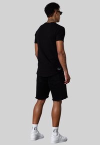 T-shirt noir à manches courtes, associé à un short en jean noir. Le modèle porte des baskets blanches et des chaussettes blanches visibles ; détail de logo simple sur le shirt.