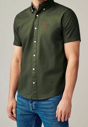 Homme portant une chemise à manches courtes couleur vert olive avec un logo rouge brodé et un jean bleu, debout avec les bras détendus.