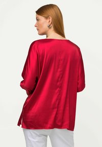 Blouse en soie rouge à coupe ample, avec des manches larges et un ourlet asymétrique, présentant une texture lisse et un léger éclat.