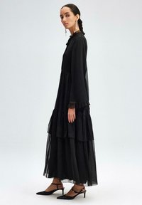 Black tiered long dress made of sheer fabric with long sleeves, ruffled cuffs, and a fitted bodice. Worn with black heeled shoes.