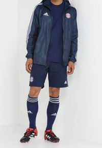 Marinblå adidas träningsdräkt med vita ränder, logga på bröstet, matchande shorts, knähöga strumpor med ränder och röda fotbollsskor.