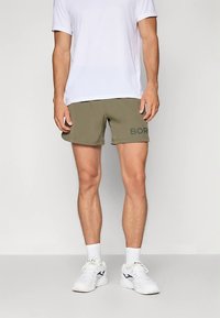 Calções verde-oliva com a palavra "BORN" em impressão escura, de corte descontraído, combinados com uma t-shirt branca lisa e ténis brancos desportivos. Textura de tecido suave.