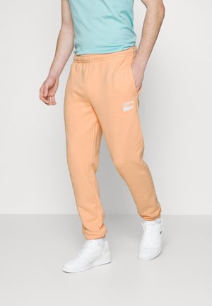 Person iført ferskenfarvede Lacoste joggingbukser, lyseblå t-shirt og hvide sneakers, stående mod en ensfarvet lys baggrund.