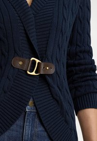 Tmavomodrý pletený kardigán s vrkočovým vzorom, sohnedým koženým opaskom so zlatou prackou v páse a rebrovanými rukávmi.