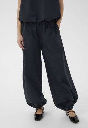 Mujer con pantalones negros sueltos con puños elásticos y un top negro a juego, combinados con zapatos negros brillantes estilo Mary Jane.