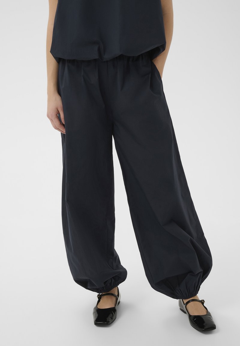 Femme portant un pantalon noir ample avec des poignets élastiques et un haut noir assorti, associée à des chaussures Mary Jane noires brillantes.