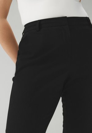 Lieknos juodos kelnės su diržo kilpomis, dėvimos su įkištu balto viršutinio drabužio, rodoma nuo vidurio liemens iki viršutinės šlaunų dalies.