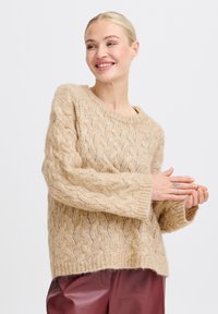 Vaaleanbeige palmikkoneule, jossa on leveä kaula-aukko ja pitkät hihat, yhdistettynä viininpunaisiin nahkahousuihin. Pehmeä, ympäristöystävällinen villamateriaali.