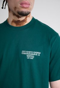 Grønn bomull T-skjorte med rund hals; har hvit brodert "Carhartt WIP"-logo på venstre brystområde. Myk tekstur, avslappet passform.