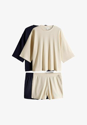 To sæt ribstrikket afslapningstøj med oversize toppe og shorts i creme og sort farve, lagt fladt på en hvid baggrund.