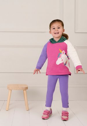 Růžový a fialový dlouhý rukáv s designem jednorožce, v kombinaci s fialovými legínami a růžovými botami na suchý zip, stojící vedle dřevěného stolu.