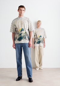 Overdimensionerede beige grafiske t-shirts med naturmotiver, parret med blå denim jeans og cremebukser. Glat tekstur, afslappet pasform.
