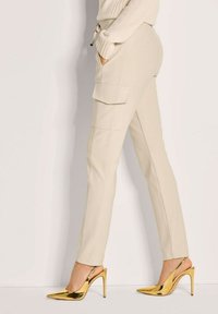 Vrouw in beige cargobroek en gouden hoge slingback-hakken, staand tegen een effen witte achtergrond.
