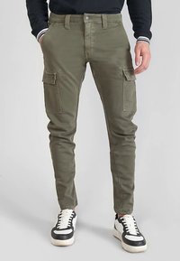 Pantalones cargo de color verde oliva hechos de mezcla de algodón, con bolsillos laterales, diseño ajustado y cierre con botón metálico. Zapatillas negras y blancas.