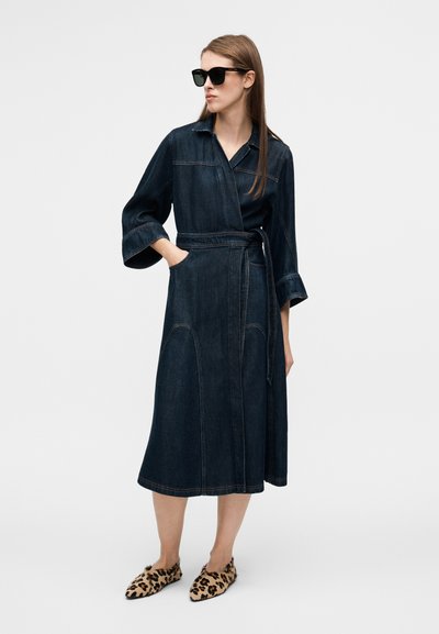 Robe portefeuille en denim bleu foncé, avec col, manches 3/4, ceinture à la taille et grandes poches. Portée avec des chaussures à imprimé léopard et des lunettes de soleil.