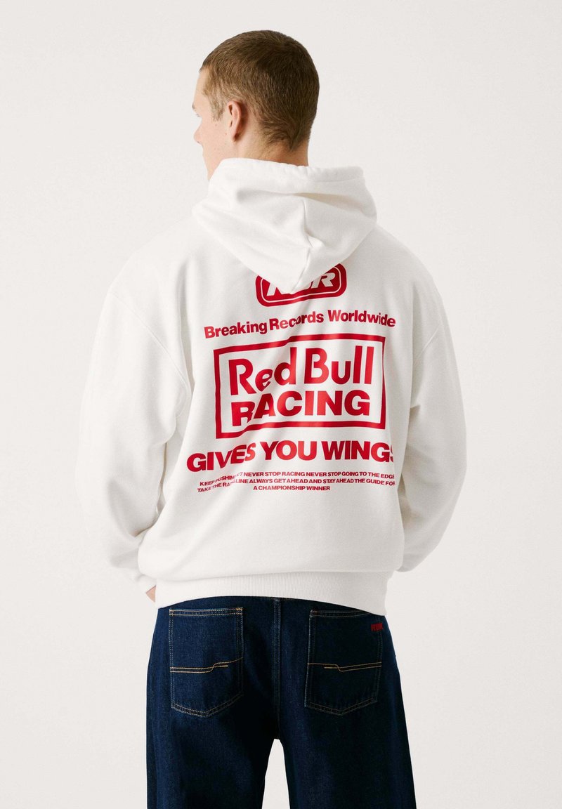 Sudadera blanca con capucha y texto rojo en la parte trasera, que presenta la frase "¡Red Bull Racing TE DA ALAS!" y detalles adicionales de la marca.