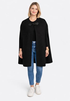 Frau trägt einen schwarzen Umhangmantel mit Verschluss, blaue Jeans, weiße Sneaker und ein schwarzes langärmeliges Oberteil, steht vor einem weißen Hintergrund.