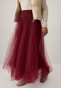Vīna sarkana tilla svārki ar plūstošu dizainu, caurspīdīgu faktūru un savilktu jostu, kopā ar bēša krāsas pūkaino vestu un gaišām kurpēm.