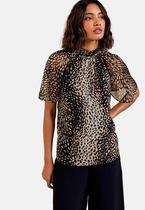 Woman with shoulder-length wavy hair wearing a black and beige spotted sheer blouse with flutter sleeves and black pants.