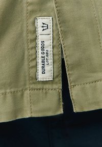 Etikett auf einem khakifarbenen Stoffkleidungsstück, mit weißem Text "DURABLE GOODS" und "LOT 082" sowie einem einfachen Kronensymbol über dem Text.