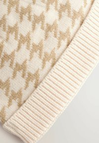 Stickad mössa med hounds-tooth mönster i beige och kräm. Har en ribbad krämfärgad mudd nederst som visar ett mjukt, texturerat material.