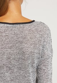 Haut en tricot gris avec un décolleté large, un tissu texturé et une bordure noire le long du décolleté, présentant un léger motif chiné.