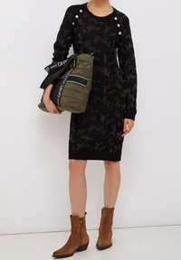 Svart stickad klänning med en strukturerad mönster, utrustad med silverknappar på axlarna. Matchas med en grön puffermapp och bruna ankelstövlar.