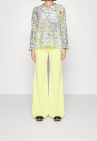 Transparente, mehrfarbige Bluse mit langen Ärmeln und V-Ausschnitt, kombiniert mit hoch taillierten, weit geschnittenen Neon-gelben Hosen. Schwarze Clogs mit goldenen Akzenten.