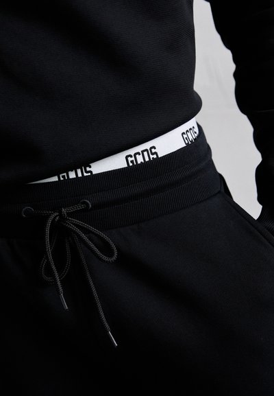 Pantalones deportivos negros con cintura ajustable, cuentan con una cinturilla elástica blanca con la marca "GCOS". Textura suave y ajuste ajustado.