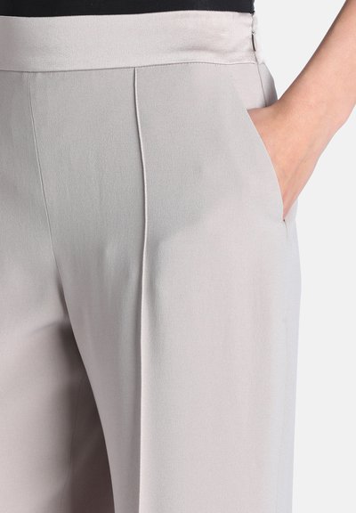 Gros plan d'un pantalon gris clair avec une poche latérale, comportant une main partiellement dans la poche et un détail de pli sur le devant.