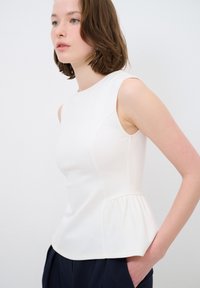Weißes ärmelloses Oberteil mit Peplum-Design, glatter Textur und Nahtdetails. Getragen mit marineblauen Hosen, vor einem neutralen Hintergrund präsentiert.