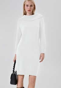 Abito bianco a maglia con un ampio scollo a cowl, maniche lunghe e orlo a coste. Indossato con una borsa nera a manico intrecciato. Texture liscia.