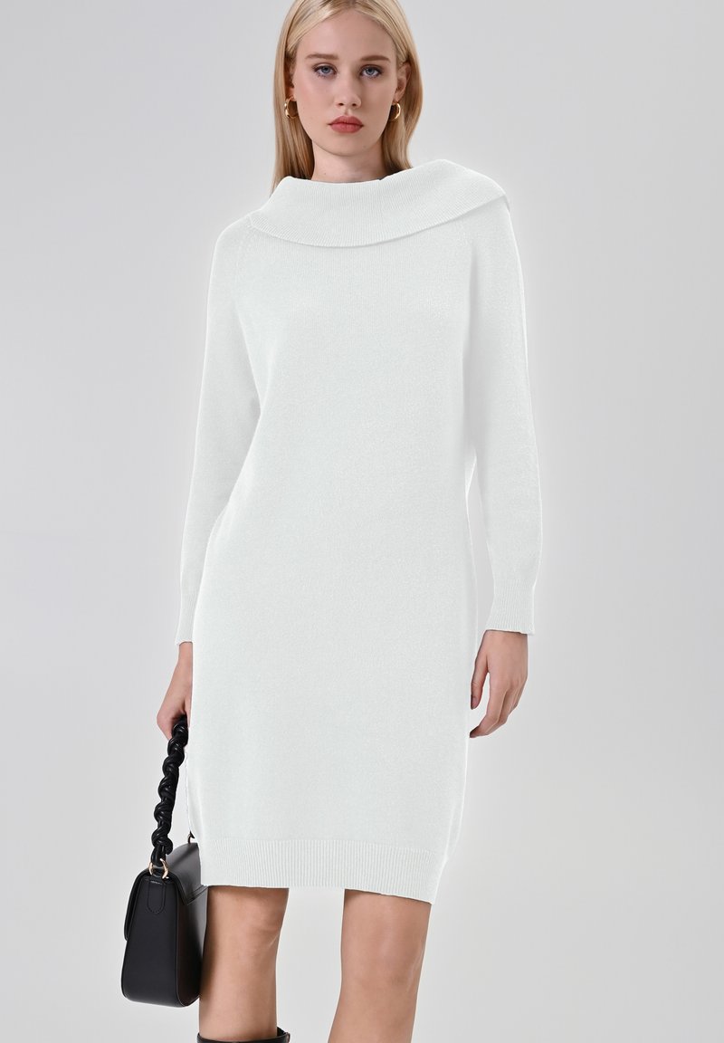 Abito bianco a maglia con un ampio scollo a cowl, maniche lunghe e orlo a coste. Indossato con una borsa nera a manico intrecciato. Texture liscia.