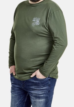 Mężczyzna w oliwkowozielonej koszuli z długim rękawem z napisem "Wszystkie dobre rzeczy są dzikie i wolne" oraz w niebieskich dżinsach, z rękami w kieszeniach, na białym tle.