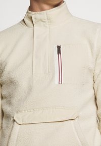 Pullover in pile crema con colletto alto, tasca frontale con zip e tasca a marsupio inferiore. Presenta dettagli con zip a contrasto.