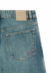 Bakfickan på blå denimjeans med synlig sömnad, en tan läderlapp på midjebandet och en liten broderad logotyp i fickans hörn.