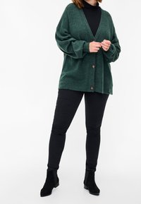 Grüne gestrickte Strickjacke mit lockerem Schnitt, tiefem V-Ausschnitt und drei großen Knöpfen. Kombiniert mit schwarzen engen Jeans und Stiefeletten.