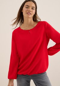 Röd blus med rund halsringning, knappstängning fram, långa ärmar och avslappnad passform. Slät tyg med mjuka draperingar.