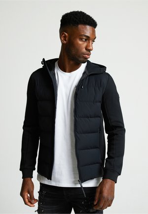 Schwarze gepolsterte Jacke mit Reißverschluss vorne und Kapuze. Sie hat Fleecefutterärmel, gerippte Bündchen und Seitentaschen. Getragen über einem weißen T-Shirt.