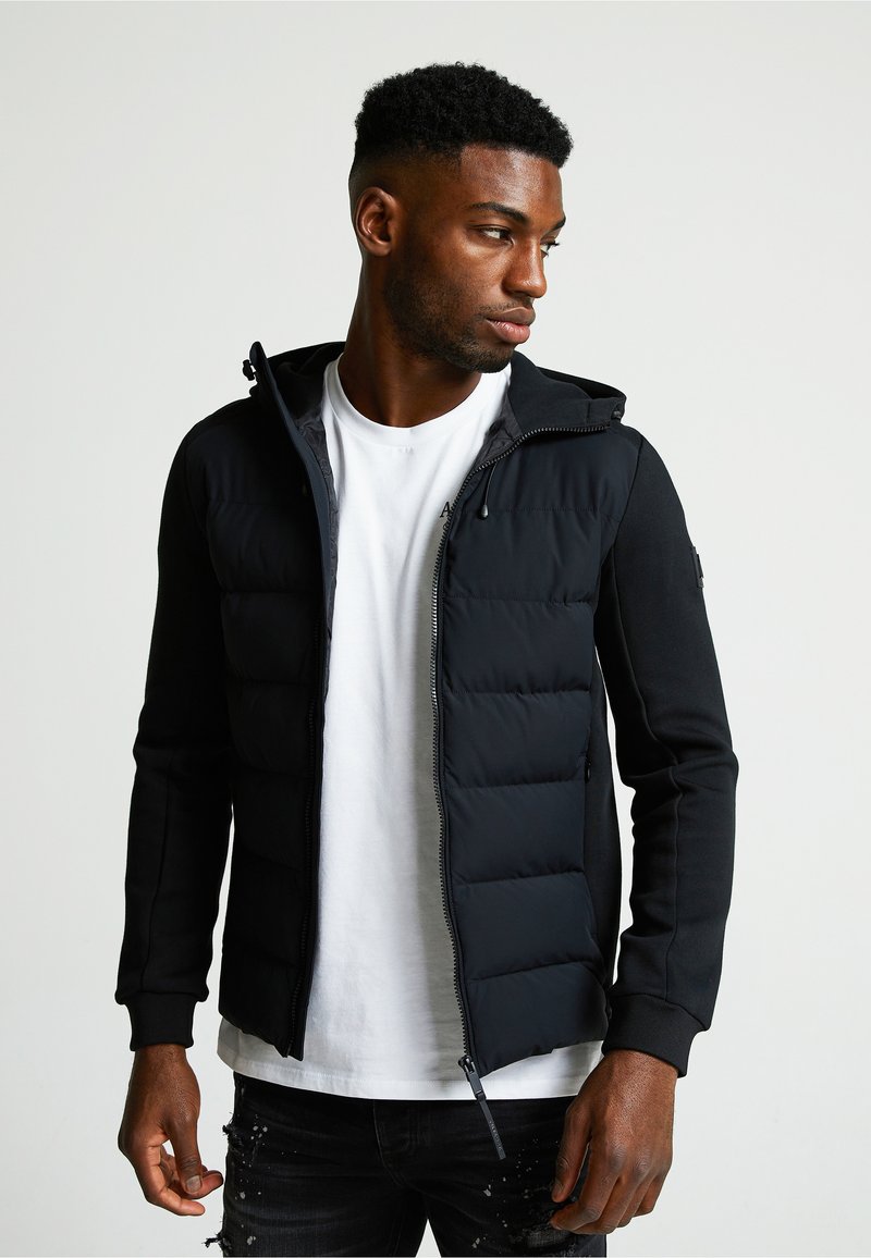 Crna jakna s podstavom, s patentnim zatvaračem sprijeda i kapuljačom. Ima flis rukave, rebraste manžete i bočne džepove. Nosena preko bijele majice.