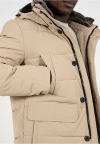 Giacca a vento beige con zip anteriore e bottoni a scatto, dotata di cappuccio con fodera in pelliccia grigia e tasche laterali. Tessuto liscio e isolato.