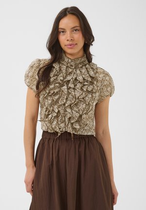 Woman with long dark hair wearing a beige floral ruffled blouse with puff sleeves and a high neck, paired with a high-waisted brown skirt.