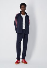 Chándal deportivo en color azul marino con franjas rojas y blancas, que incluye una chaqueta con cremallera y pantalones con cintura elástica, combinado con zapatillas rojas.