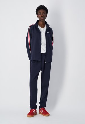 Tuta sportiva navy con strisce rosse e bianche, composta da giacca con zip e pantaloni con vita elastica, abbinata a sneakers rosse.