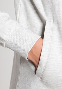 Sudadera gris con una tela suave y texturizada. Presenta un puño acanalado y un bolsillo lateral, mostrando un diseño simple y funcional.