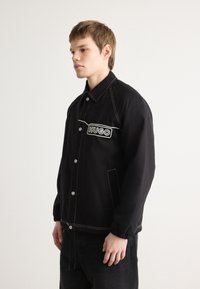 Giacca nera in tessuto, con chiusura a bottoni, cuciture bianche e un patch con logo. Include tasche laterali e polsini elasticizzati.