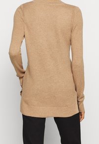 Vue de dos d'une personne portant un pull en tricot texturé beige à manches longues et un pantalon noir, debout devant un fond blanc uni.