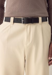 Cinturón de cuero negro con una hebilla plateada, llevado sobre pantalones beige de textura suave y delantero plisado. Suéter marrón visible por encima.