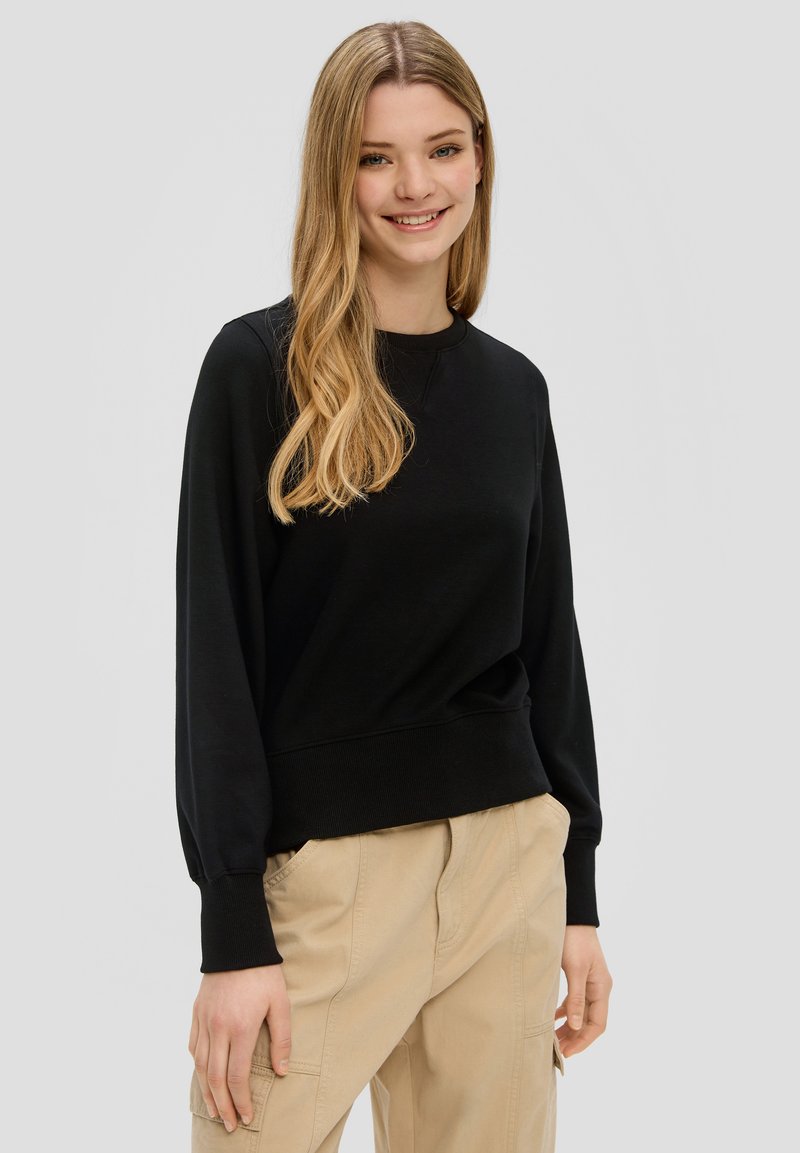 Zwarte sweatshirt met lange mouwen, geribbelde ronde halslijn en manchetten. Zacht materiaal, comfortabele pasvorm, gecombineerd met lichtbeige broek.